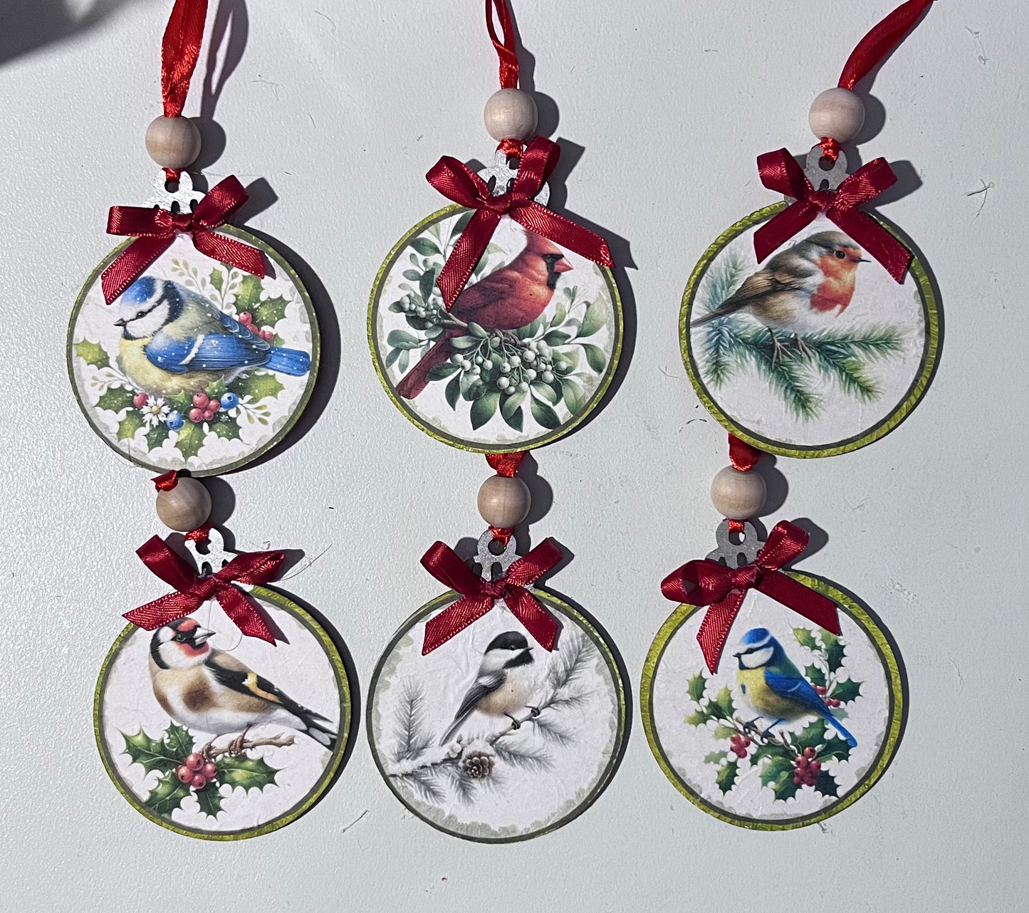 Set of six winter bird Christmas ornaments on wooden rounds with cardinals, chickadees, and woodland birds surrounded by pine greenery, berries, and winter foliage, finished with red ribbons, gingham bows, and wood beads.