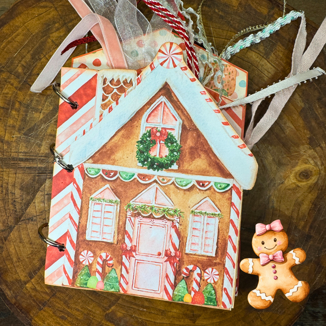Close-up of the "Gingerbread Dreams" Christmas junk journal folio. The cover features a gingerbread house decorated with candy canes, peppermint swirls, and a holiday wreath. Ribbons are tied at the top, and a small smiling gingerbread girl charm with a pink bow is placed next to the journal.