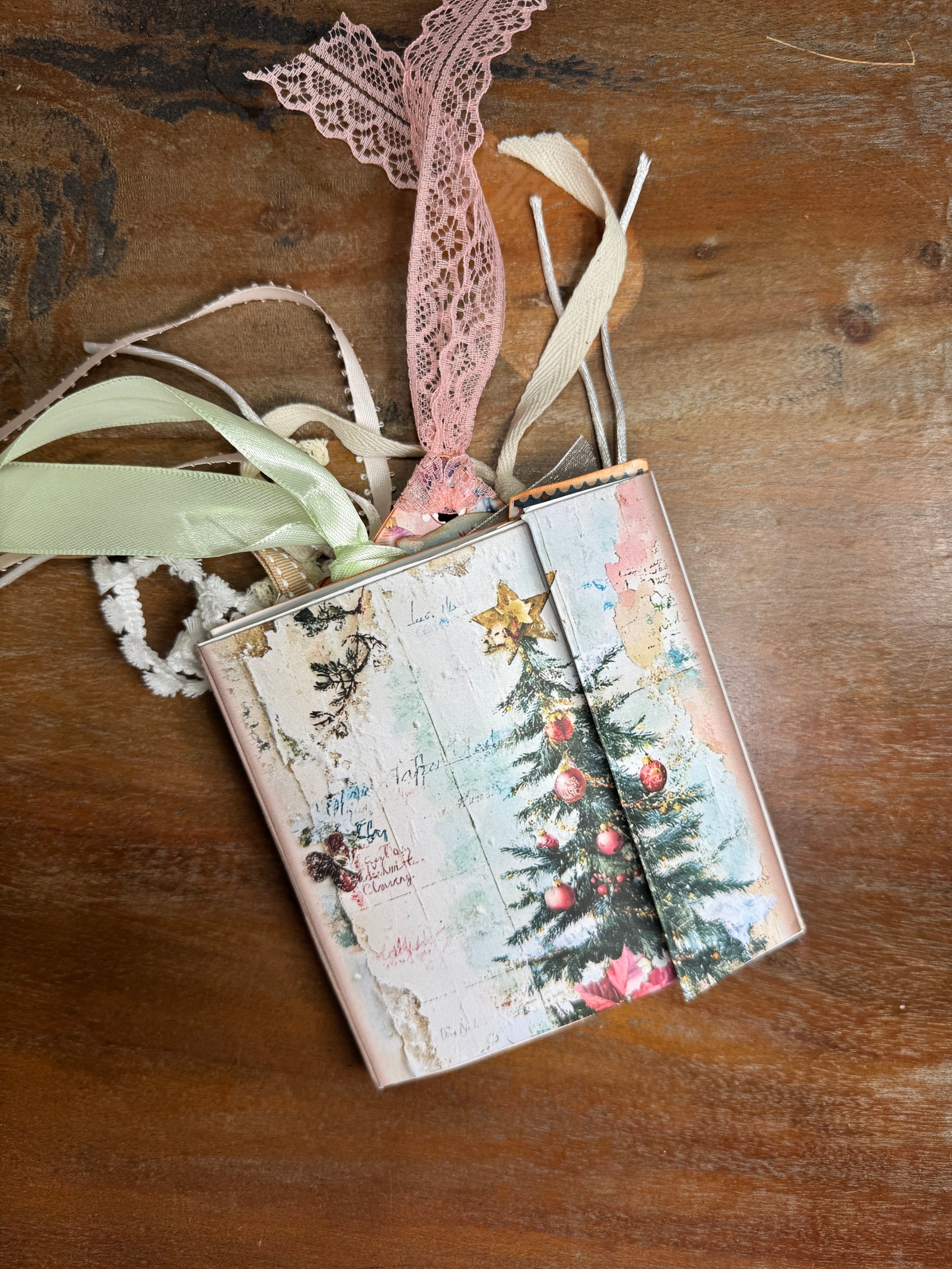 Handmade Nutcracker Christmas junk journal folio with a vintage-style cover featuring a decorated Christmas tree with red ornaments and a golden star. The journal is tied with pastel ribbons and lace in pink, cream, and green, spilling from the top. It rests on a wooden surface.