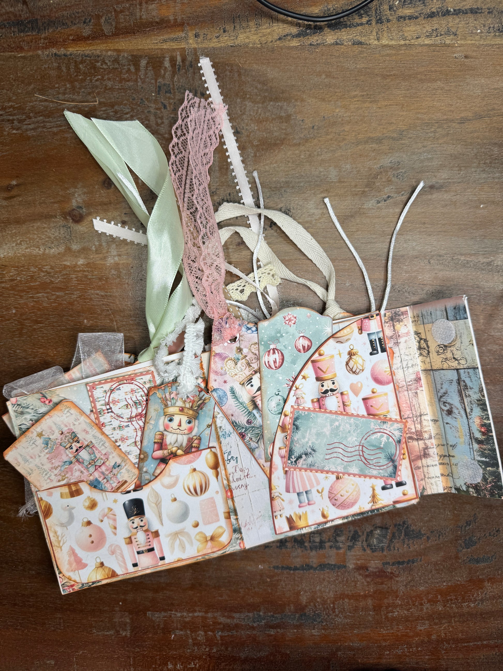 Handmade Nutcracker-themed Christmas junk journal folio opened to decorated pages. The spread includes pastel holiday papers with nutcracker soldiers, ballerinas, ornaments, and festive patterns. Pockets hold tags and journaling cards, while pastel ribbons and lace in pink, cream, and green extend from the top of the journal.