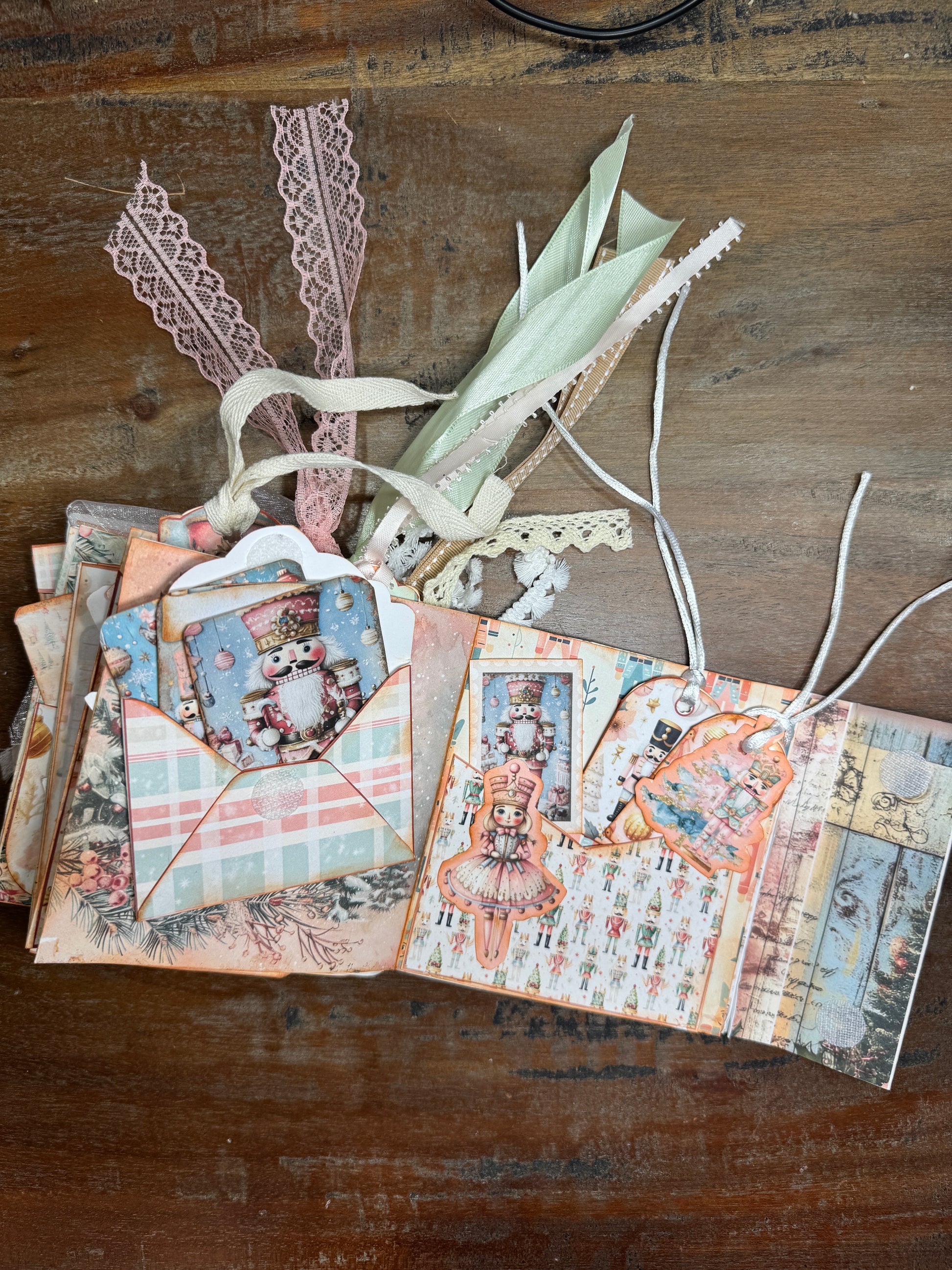 Open Nutcracker-themed Christmas junk journal folio displayed on a wooden surface. Inside pages feature pastel holiday designs with nutcracker soldiers, a ballerina, patterned paper, and tuck spots holding tags and inserts. Ribbons and lace in pink, cream, and green extend from the top of the journal.