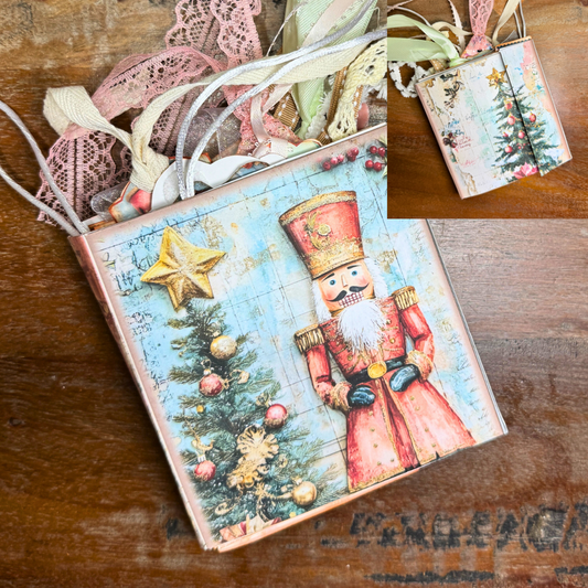 Close-up of a handmade Nutcracker Christmas junk journal folio. The cover features a vintage-style Nutcracker soldier in red next to a decorated Christmas tree topped with a golden star. Ribbons, lace, and tags peek from the top of the journal. A smaller inset image shows the journal closed with both front panels decorated with Christmas trees.