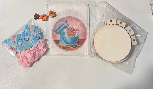 Christmas Bake Shop ornament craft kit featuring seven pastel baking-themed ornaments displayed on a flat surface, including cupcakes, gingerbread cookie scenes, retro mixers, rolling pin dough, stacked bowls, pink kitchen scenes, and a polka-dot apron, decorated with bows, fabric ties, and gingerbread embellishments.