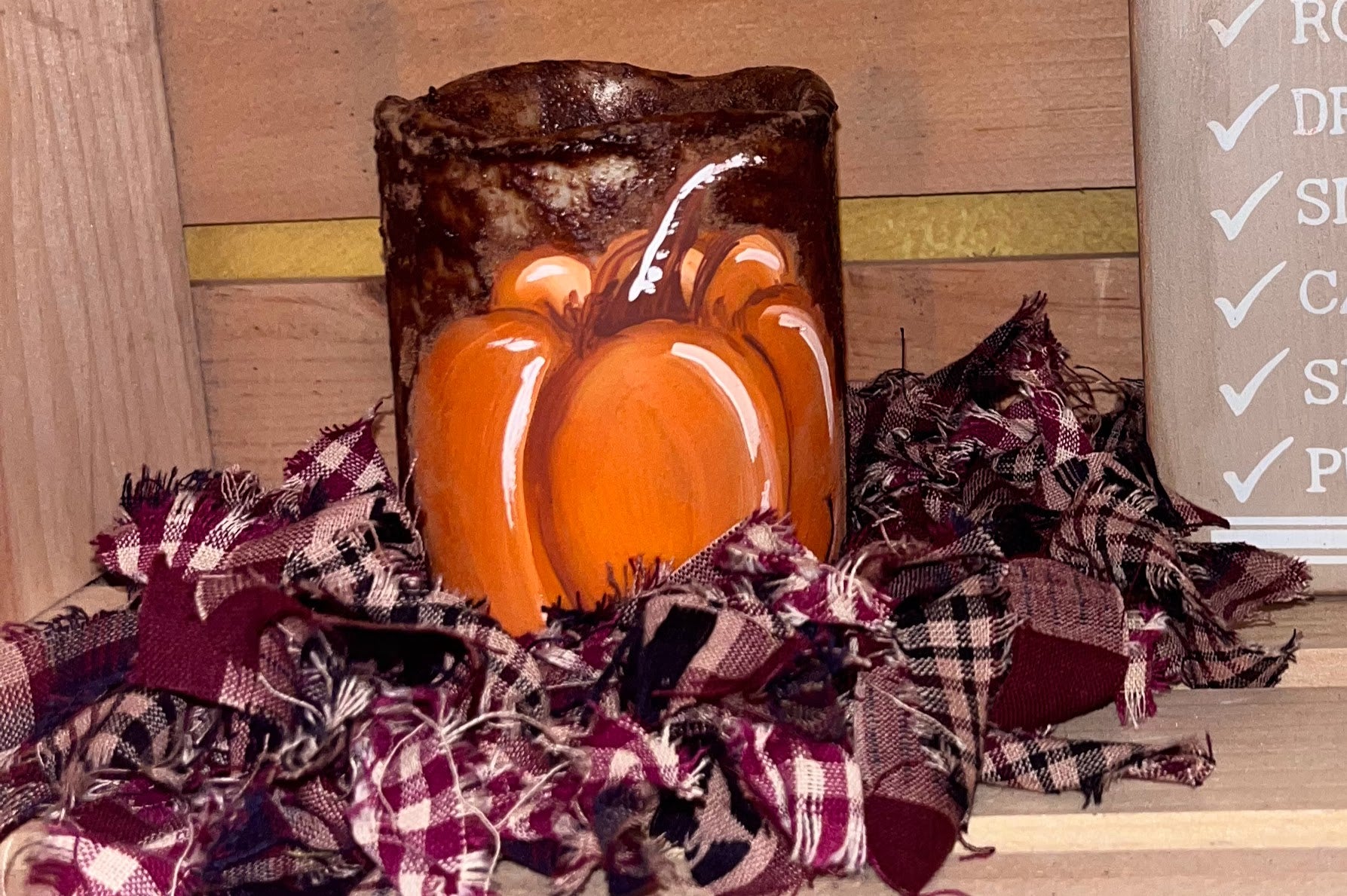 Hand-painted primitive pumpkin flameless candle with a cinnamon-grunge finish, surrounded by burgundy and cream plaid rag garland, styled in a rustic fall decor setting.