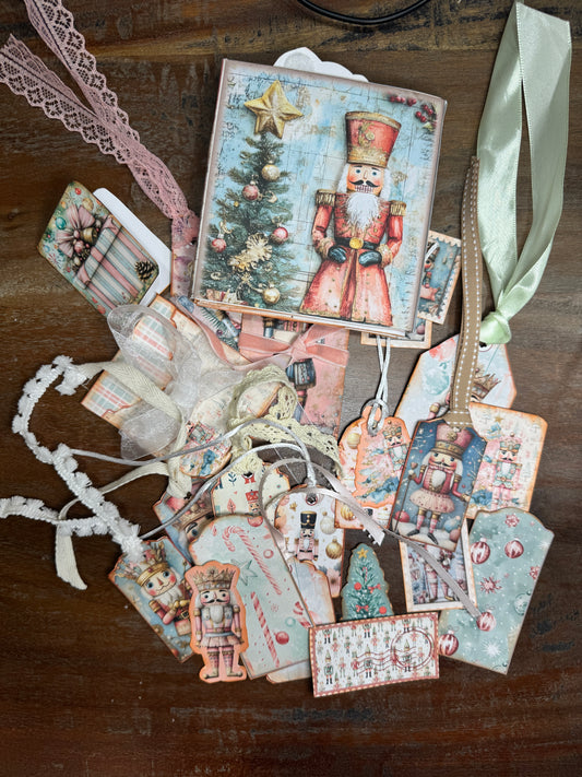 Handmade Nutcracker-themed junk journal folio displayed with an assortment of coordinating tags and inserts. The cover shows a red Nutcracker soldier beside a Christmas tree. Around it are various decorated tags featuring nutcrackers, ballerinas, candy canes, ornaments, and holiday patterns, each tied with ribbon or lace.