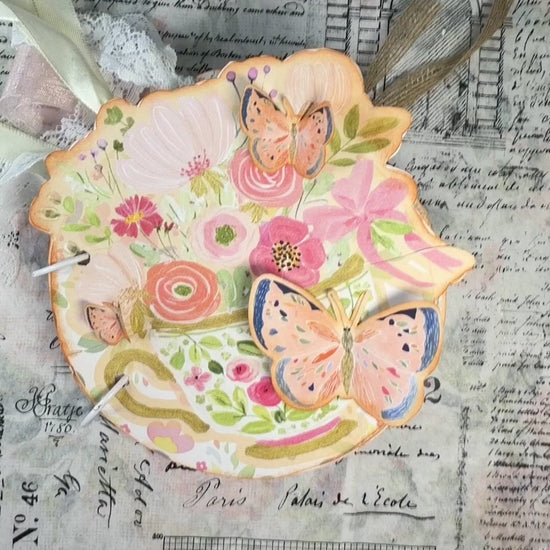 Hands flip through a floral teacup-shaped Afternoon Tea journal, revealing butterfly embellishments, pockets, tags, and ribbons.