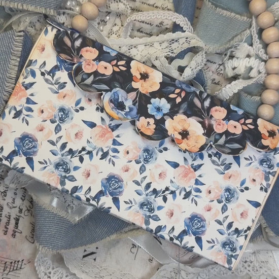 Hands open a blue floral wedding accordion journal with a church page, pockets, tags, ribbons, and photo mats.