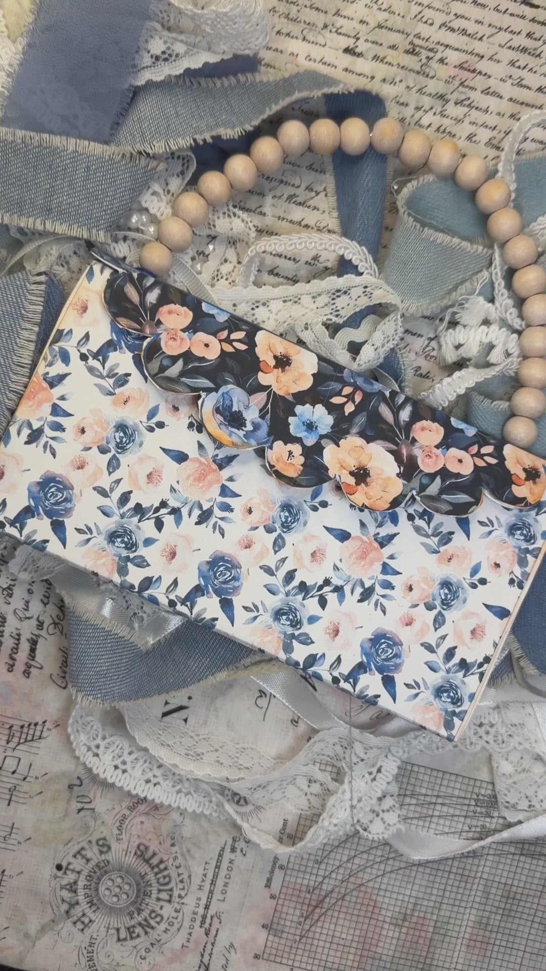 Hands open a blue floral wedding accordion journal with a church page, pockets, tags, ribbons, and photo mats.