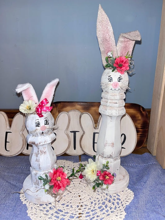 Two wooden spindle bunnies with florals at their ears and the base setting on a tabletop with a doily underneath and easter sign behind them